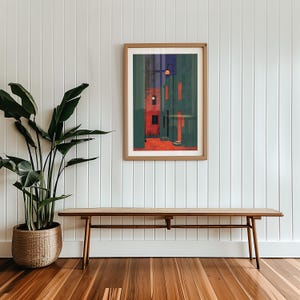以下が含まれることがあります： 赤とティール色の建物が描かれた、様式化された街並みのフレーム入りアートプリント。 アートワークは木製のフレームに入っており、白いパネルの壁に掛けられています。 ベンチと鉢植えの植物が前景にあります。