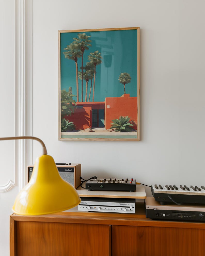 May include: A framed art print depicts a mid-century modern style building with palm trees against a teal sky. The building is a vibrant orange with a flat roof. The artwork is displayed above a wooden cabinet with a yellow lamp, audio equipment, and a keyboard.