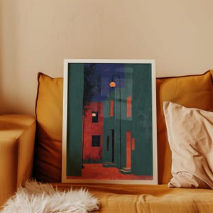 以下が含まれることがあります： 緑、赤、青の色合いの建物が描かれた抽象的な街並みのフレーム入りアートプリント。オレンジ色の光を放つ街灯がシーンを照らしています。マスタード色のソファに飾られたアートワーク。