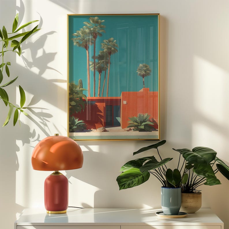 May include: An orange and red table lamp with a rounded shade and base sits on a white surface. Above it, a framed print depicts a desert scene with palm trees and a red building against a teal sky. Green plants are in the foreground.