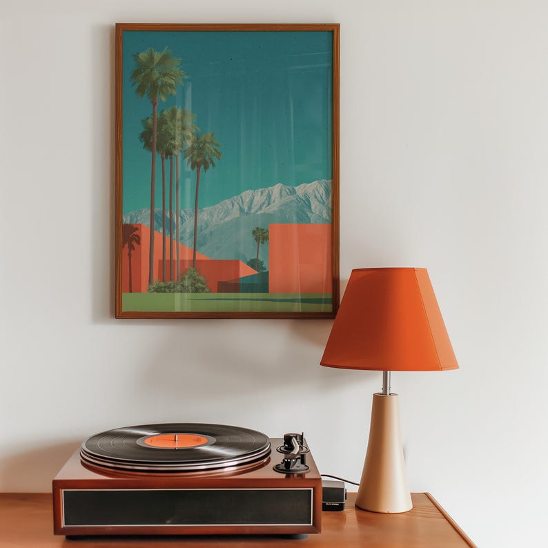 May include: A framed art print depicting a desert landscape with palm trees, red buildings, and mountains. A vintage record player with a black vinyl record and an orange label sits below the artwork. An orange lampshade sits atop a beige lamp base.