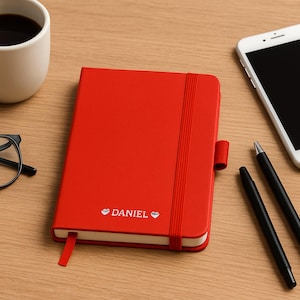 Puede incluir: Una libreta roja con cierre de banda elástica blanca y el nombre "DANIEL" impreso en la portada. La libreta está sobre una mesa de madera con un teléfono inteligente blanco, dos bolígrafos negros, una taza de café y unas gafas negras.