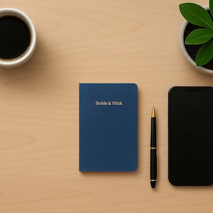 Könnte beinhalten: Ein blaues Notizbuch mit dem Text "Stride & Fitch" auf dem Cover, ein goldener Stift, ein schwarzes Smartphone und eine Topfpflanze auf einer hellen Holzoberfläche.