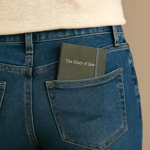 May include: A dark green journal with the text "The Diary of Jane" on the front cover is tucked into the back pocket of a pair of blue jeans.