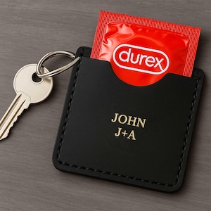 Może przedstawiać: Czarny skórzany brelok do kluczy z imieniem "JOHN" i inicjałami "J+A" w złotych literach. Czerwona prezerwatywa Durex jest częściowo widoczna w kieszeni. Do breloka przymocowany jest srebrny klucz.