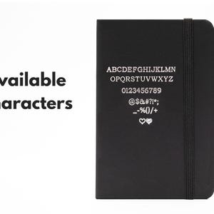 May include: A black notebook with a silver inscription listing available characters for personalization. The inscription includes the alphabet, numbers 0-9, and symbols such as @, $, #, %, and hearts.