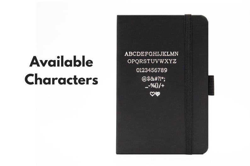 May include: A black notebook with a silver inscription listing available characters for personalization. The inscription includes the alphabet, numbers 0-9, and symbols such as @, $, #, %, and hearts.