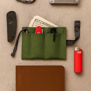 May include: An assortment of everyday carry items, including a green canvas wallet with cash, a Leatherman multi-tool, a black folding knife, a red lighter, a carabiner, a pen, and a brown notebook. The items are arranged on a neutral surface.
