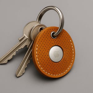 Könnte beinhalten: Ein brauner Leder-Schlüsselanhänger mit einem runden AirTag-Halter und einem silbernen Apple-Logo. Der Schlüsselanhänger hat einen genähten Rand und ein perforiertes Design.