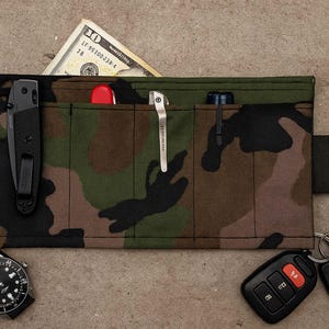 May include: Camouflage-patterned roll-up organiser with multiple pockets, holding a black knife, pen, flashlight, and a hundred-dollar bill. A black watch, car keys, and a car remote are also visible.