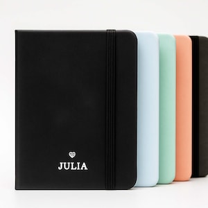 Pode incluir: Cinco cadernos em cores diferentes: preto, azul claro, verde claro, pêssego e cinza escuro. O caderno preto tem um coração prateado e o nome "JULIA" impresso na capa.