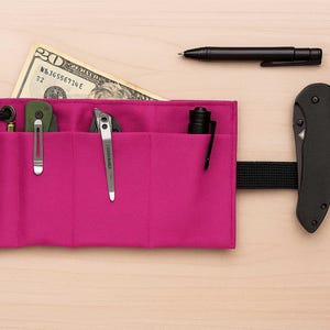 May include: A magenta roll-up organiser with various tools and accessories. Includes a black pen, a folding knife, a small torch, and a pocket knife. A twenty-dollar bill is visible. The organiser is secured with a black strap.