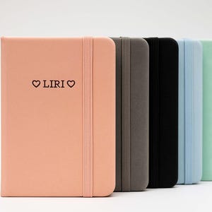 Op de afbeelding: Een set van zes notitieboekjes met verschillende gekleurde omslagen. De voorkant van het eerste notitieboekje is perzikkleurig en heeft de tekst "♡ LIRI ♡" erop gedrukt. De andere notitieboekjes zijn grijs, zwart, lichtblauw en lichtgroen.