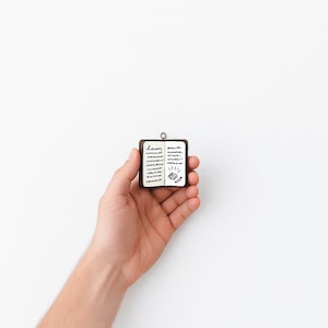 May include: A small, brown, leather-bound book with a white page open to reveal handwritten notes and a small drawing of a lightbulb. The book is being held in a person's hand.