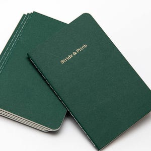 Op de afbeelding: Een stapel donkergroene notitieboekjes met gouden stiksels en de tekst "Stride & Pitch" op het bovenste notitieboekje.