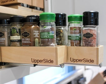 May include: A close-up of a wooden spice rack with various spices in glass jars. The jars are labeled with the names of the spices, such as "onion powder", "cardamom", "celery salt", "six chili powder", "allspice", "cinnamon sticks", and "sea salt". The rack is labeled "UpperSlide" on the front.