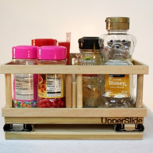 May include: A wooden slide-out shelf with a label that reads "UnderSlide". The shelf is filled with various glass jars and bottles, including pink jars with colorful sprinkles, a red jar with red candies, a clear jar with honey, and a clear bottle with a bear-shaped design.