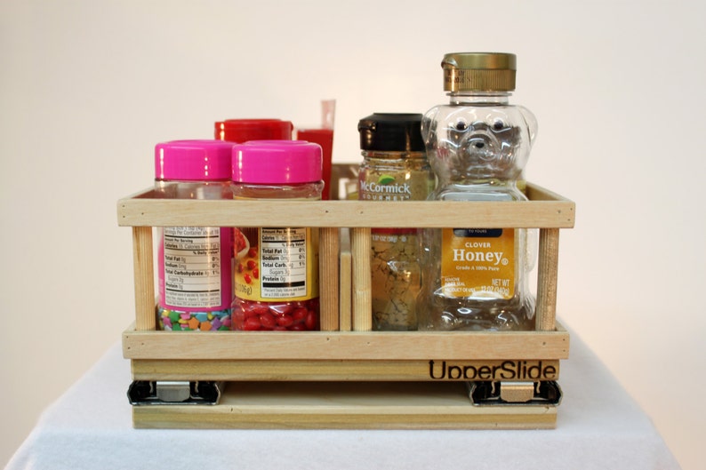 May include: A wooden slide-out shelf with a label that reads "UnderSlide". The shelf is filled with various glass jars and bottles, including pink jars with colorful sprinkles, a red jar with red candies, a clear jar with honey, and a clear bottle with a bear-shaped design.