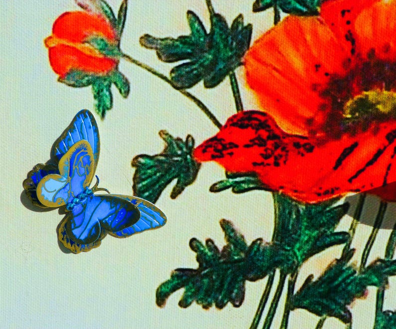 Puede incluir: Un broche de mariposa azul con bordes dorados y patrones de alas intrincados, colocado junto a una ilustraci&oacute;n impresa de vibrantes amapolas rojas con hojas de color verde oscuro sobre un fondo de color crema claro.