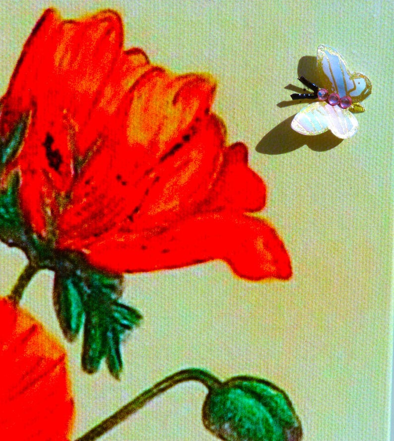 Puede incluir: Un lienzo impreso que muestra una amapola roja vibrante con hojas y tallo verdes, acompa&ntilde;ada de un peque&ntilde;o adorno de mariposa tridimensional con alas blancas y detalles de cuentas rosas situado a la derecha de la flor.