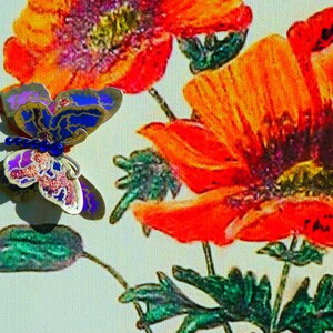 Puede incluir: Un broche de mariposa con alas met&aacute;licas azules, moradas y plateadas con una hilera de peque&ntilde;as cuentas azules en el cuerpo, colocado sobre un lienzo impreso de amapolas de color naranja y rojo vibrante con tallos y hojas verdes.