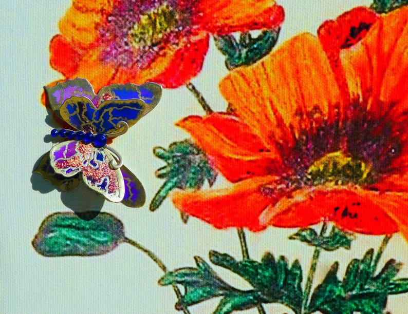 Puede incluir: Un broche de mariposa con alas met&aacute;licas azules, moradas y plateadas con una hilera de peque&ntilde;as cuentas azules en el cuerpo, colocado sobre un lienzo impreso de amapolas de color naranja y rojo vibrante con tallos y hojas verdes.
