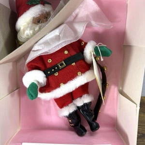 Puede incluir: Una muñeca de Papá Noel en una caja. La muñeca lleva un traje rojo con ribetes blancos, un cinturón negro y botas negras. El sombrero de la muñeca es rojo con ribetes blancos y una hoja de acebo verde. La caja es rosa y tiene papel de seda blanco.