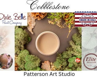 May include: A close-up of a small jar of light brown paint with the words "Cobblestone" and "Patterson Art Studio" written on the image. The jar is surrounded by green moss and pine cones. The image also includes the logos for Dixie Belle Paint Company and Elite Retailer.