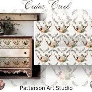 May include: A white dresser with three drawers decorated with a floral and antler design. The dresser is labelled "Cedar Creek" and is sitting in front of a white wall. The background is a repeating pattern of antlers, flowers, and greenery.