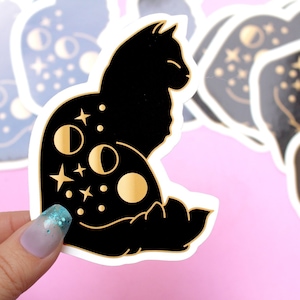 May include: A black cat silhouette sticker with gold moon and star details. The sticker is on a pink background.