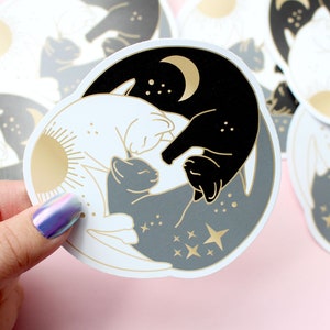 Pu&ograve; includere: Adesivi rotondi con un design yin-yang con gatti. Un lato &egrave; nero con una luna crescente, l'altro bianco con un sole. Un gatto grigio &egrave; al centro, con stelle e dettagli dorati. Gli adesivi sono tenuti da una mano.