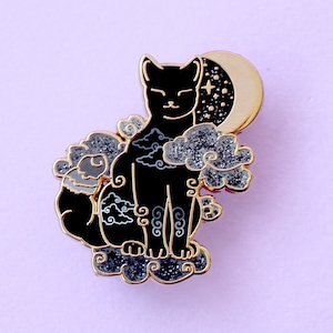 May include: Black enamel pin of a cat with a crescent moon and stars on its back. The cat is surrounded by clouds and has a gold outline.