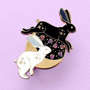 May include: A gold enamel pin featuring two rabbits, one black and one white, with a crescent moon and stars. The rabbits are stylized with floral details and a starry sky background.