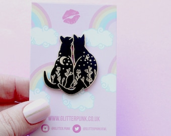 May include: Black enamel pin featuring two cats sitting back to back, with a crescent moon and stars on their bodies. The cats are surrounded by floral designs. The pin is on a pink background with a rainbow and clouds.