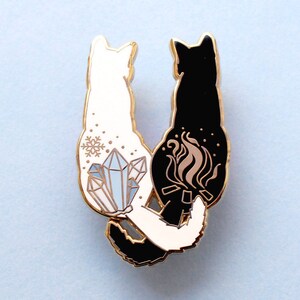 May include: Gold-tone enamel pin featuring two black cats facing each other. One cat has a snowflake and crystals on its back, the other has a flame on its back.