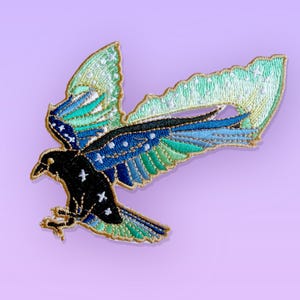 May include: An embroidered patch featuring a black raven in flight with wings spread wide. The wings display a gradient of blue, teal, and green colours with white star-shaped accents. The entire design is outlined in a metallic gold thread.