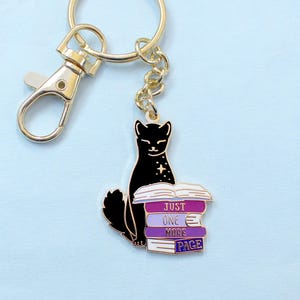 May include: A gold-toned keychain with a black cat charm sitting on a stack of books. The books are purple and white, with the text "JUST ONE MORE PAGE". The keychain has a lobster clasp and a chain.