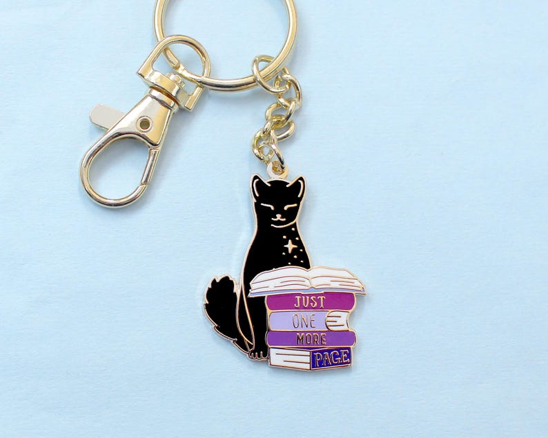 May include: A gold-toned keychain with a black cat charm sitting on a stack of books. The books are purple and white, with the text "JUST ONE MORE PAGE". The keychain has a lobster clasp and a chain.