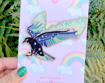 Puede incluir: Un parche bordado que muestra un pájaro negro con las alas extendidas en tonos azul verdoso, azul y verde, acentuado con motivos de estrellas blancas. El parche está montado sobre una tarjeta de soporte de color violeta claro con arcoíris.