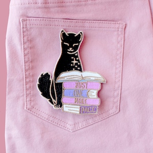 May include: A black cat patch with a white cross design sits atop a stack of books. The books are purple, blue, and white, with the words "JUST ONE MORE PAGE" embroidered on them. The patch is on a pink pocket.