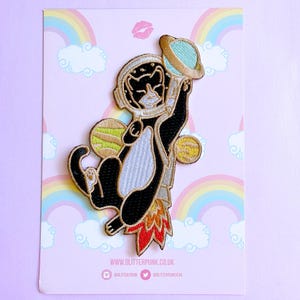 May include: An embroidered patch featuring a black and white cat wearing a space helmet and a jetpack, floating among three planets. The cat holds a light blue planet above its head. The patch is displayed on a card with a rainbow background.