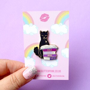 Op de afbeelding: Zwarte kat emaille pin met een stapel boeken waarop "Just One More Page" staat. De pin is op een roze achtergrond met een regenboog en wolken.