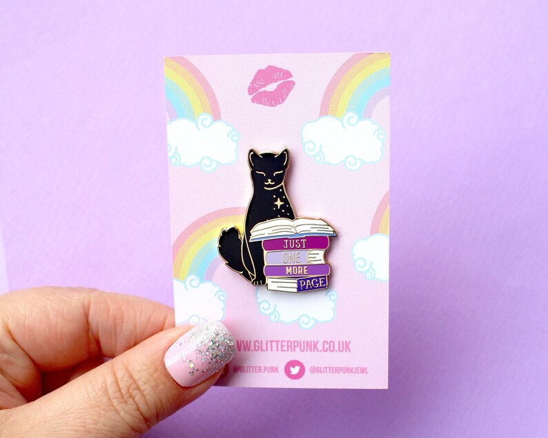 Op de afbeelding: Zwarte kat emaille pin met een stapel boeken waarop "Just One More Page" staat. De pin is op een roze achtergrond met een regenboog en wolken.