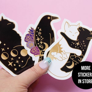 May include: Three sticker designs featuring a black cat with moon phases, a black crow with flowers, and a black and white cat hugging. The stickers have a gold foil design and are on a pink background. The text "More Stickers in Store!" is in a black circle.