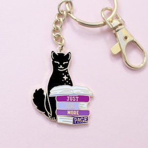 May include: A black cat keychain with a gold-colored chain and clasp. The cat is sitting next to a stack of books with the text "JUST ONE MORE PAGE" in purple and white. The background is a light pink.