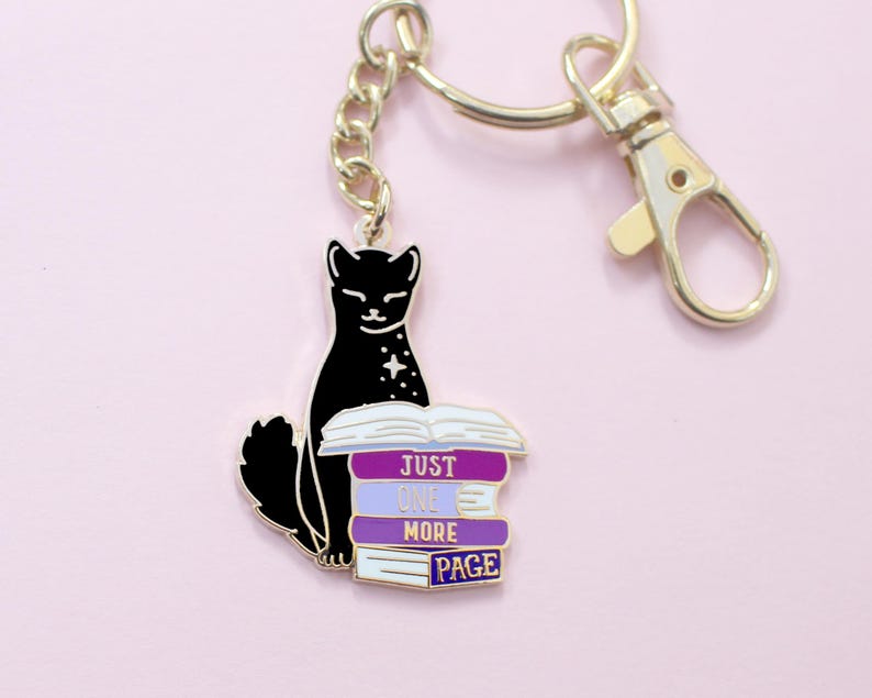 May include: A black cat keychain with a gold-colored chain and clasp. The cat is sitting next to a stack of books with the text "JUST ONE MORE PAGE" in purple and white. The background is a light pink.