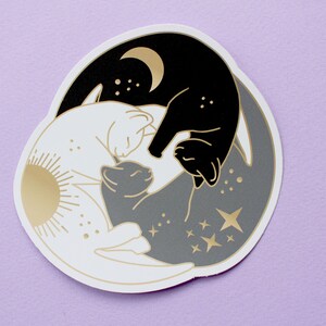 Pu&ograve; includere: Un adesivo circolare con un design yin-yang con gatti. Un gatto &egrave; nero con una luna crescente dorata, l'altro bianco con un sole dorato. Un gatto grigio al centro, con stelle e dettagli dorati.