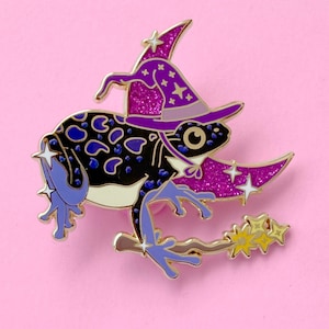 May include: A metal enamel pin featuring a black frog with blue spots wearing a purple glittery witch hat. The frog is perched on a crescent moon and holds a wand with yellow star-shaped tips. The design includes small white star accents.