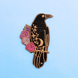 May include: Embroidered patch featuring a black raven perched on a branch, adorned with a crescent moon, stars, and floral accents in purple, pink, and gold. The patch has a gold border and is set against a light blue background.