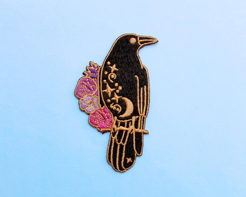 May include: Embroidered patch featuring a black raven perched on a branch, adorned with a crescent moon, stars, and floral accents in purple, pink, and gold. The patch has a gold border and is set against a light blue background.